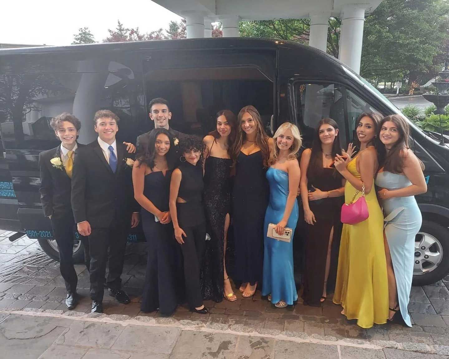 Group of people in formal attire pose in front of a black van, likely for prom.