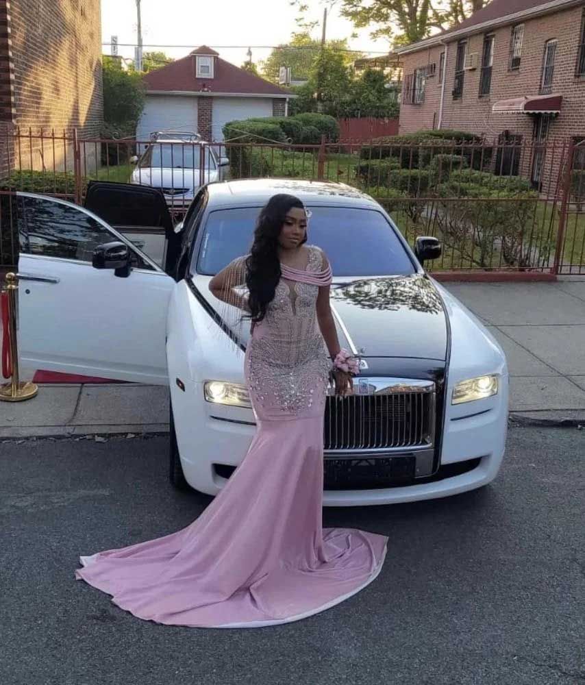 Woman in pink gown posing by a white Rolls-Royce with an open door, outside a building with a lawn.