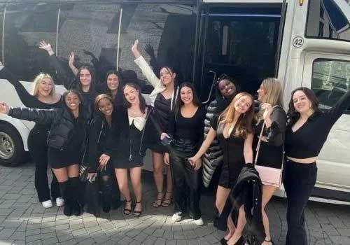 Group of women in black clothing, smiling and waving in front of a white bus.