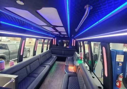 Interior of a party bus with blue and green lighting, black seating, and tinted windows.