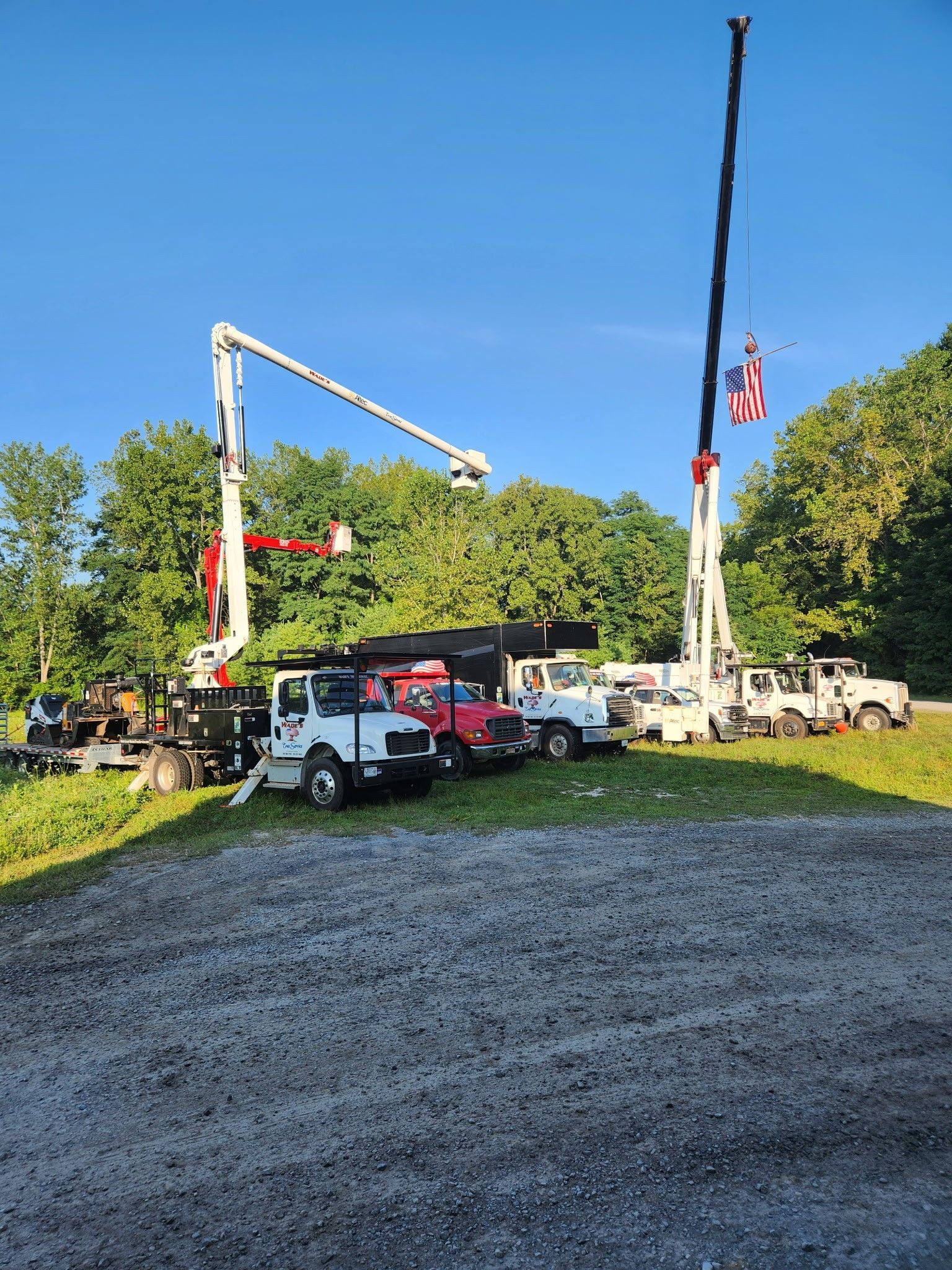 Utility trucks parked on gravel, two with extended booms against a backdrop of trees and blue sky.