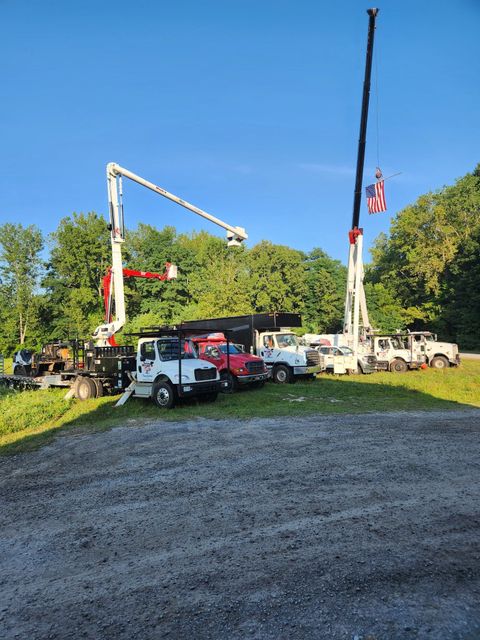 Utility trucks parked on gravel, two with extended booms against a backdrop of trees and blue sky.