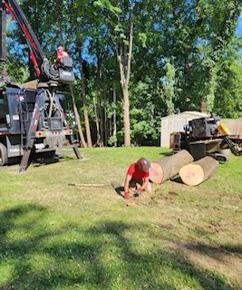 A man is kneeling down next to a pile of logs in a grassy field.