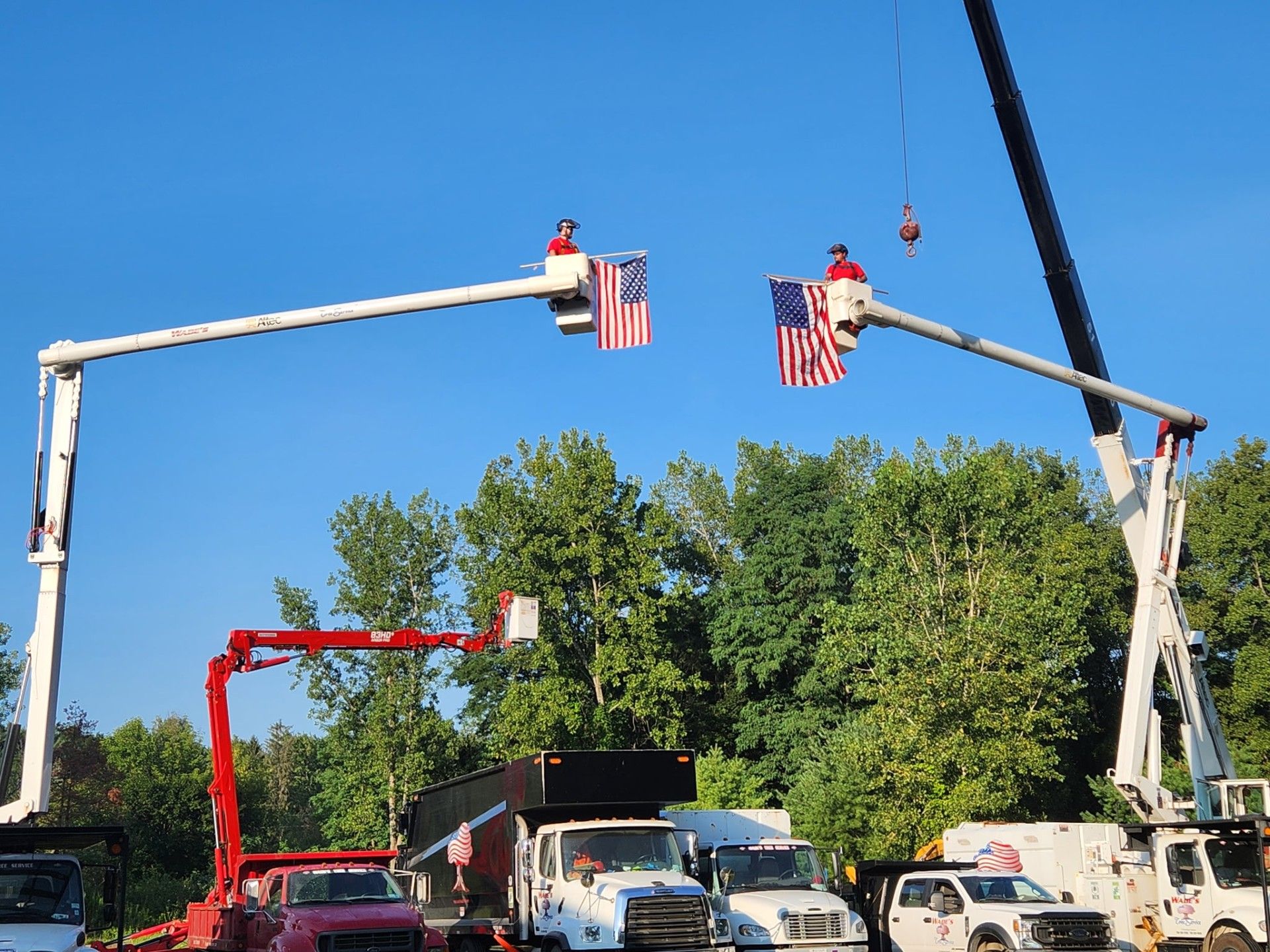 Two workers in boom lifts with American flags; trucks and trees under a blue sky.