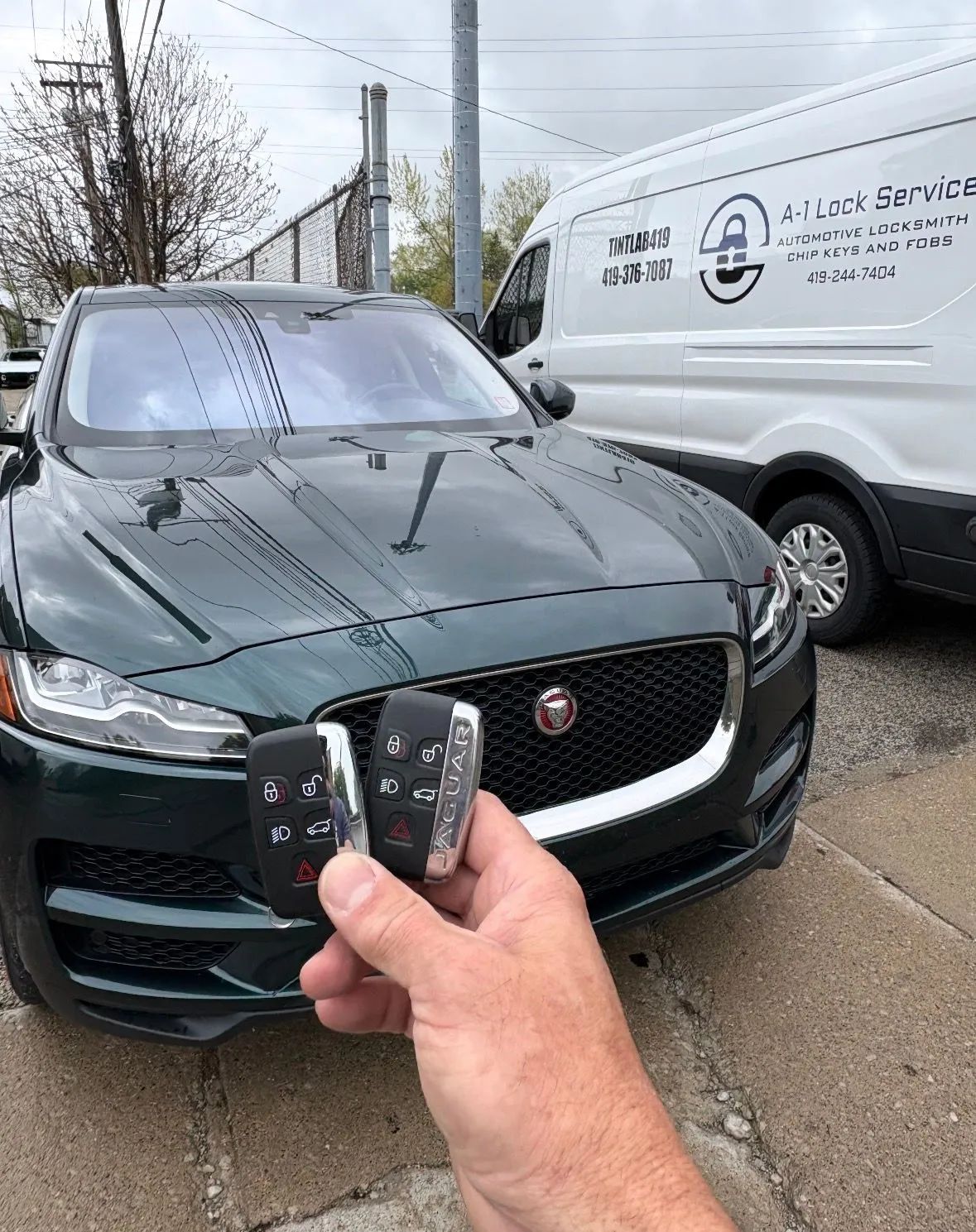Hand holding a car key fob toward a dark green sedan beside a white work van in a wet driveway