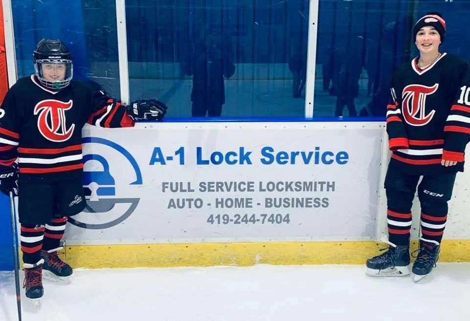 Two hockey players in black-and-red jerseys standing by an A-1 Lock Service rink board