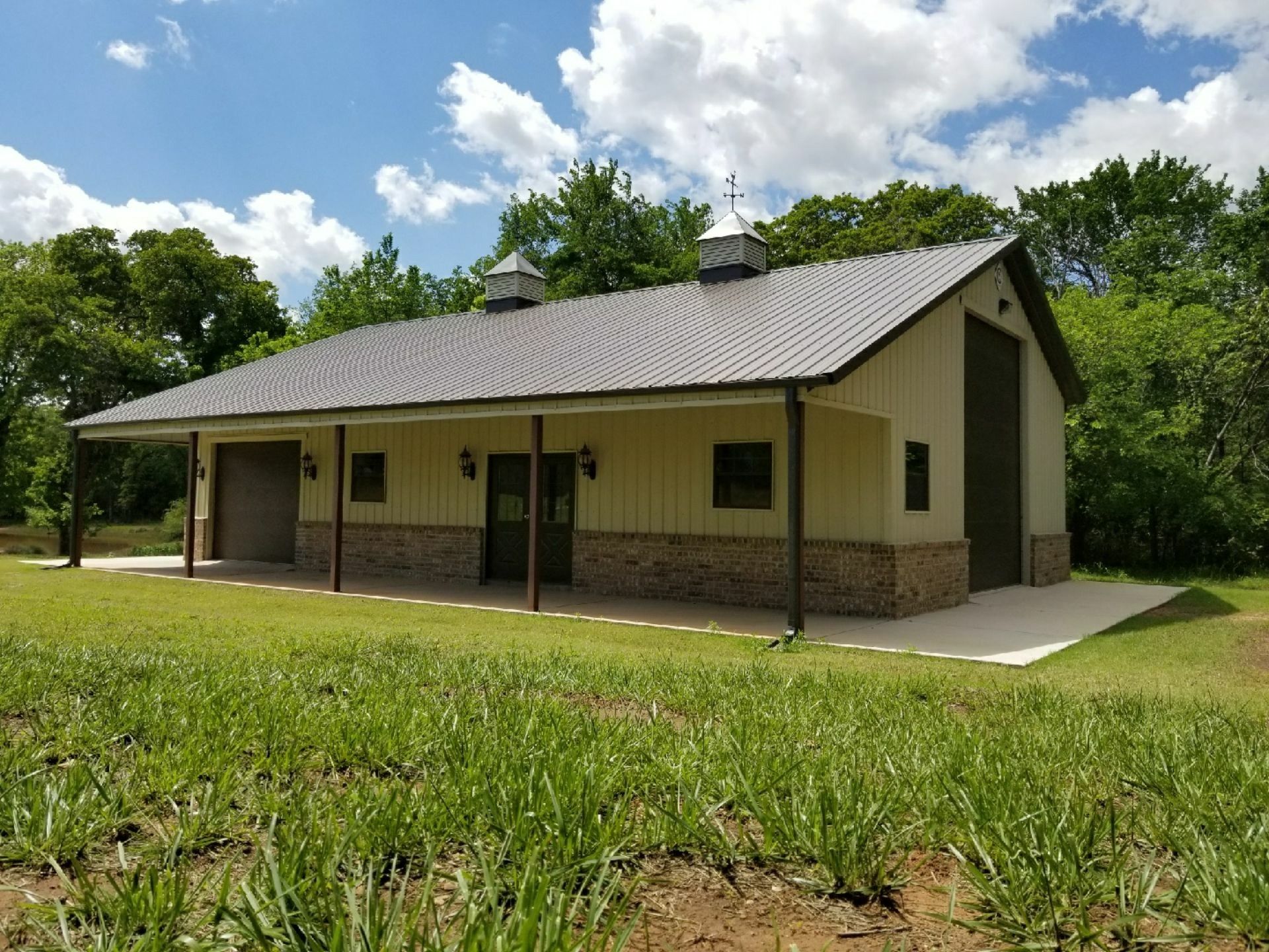 Tan barn with a porch, brown roof, and stone base, in a grassy field.