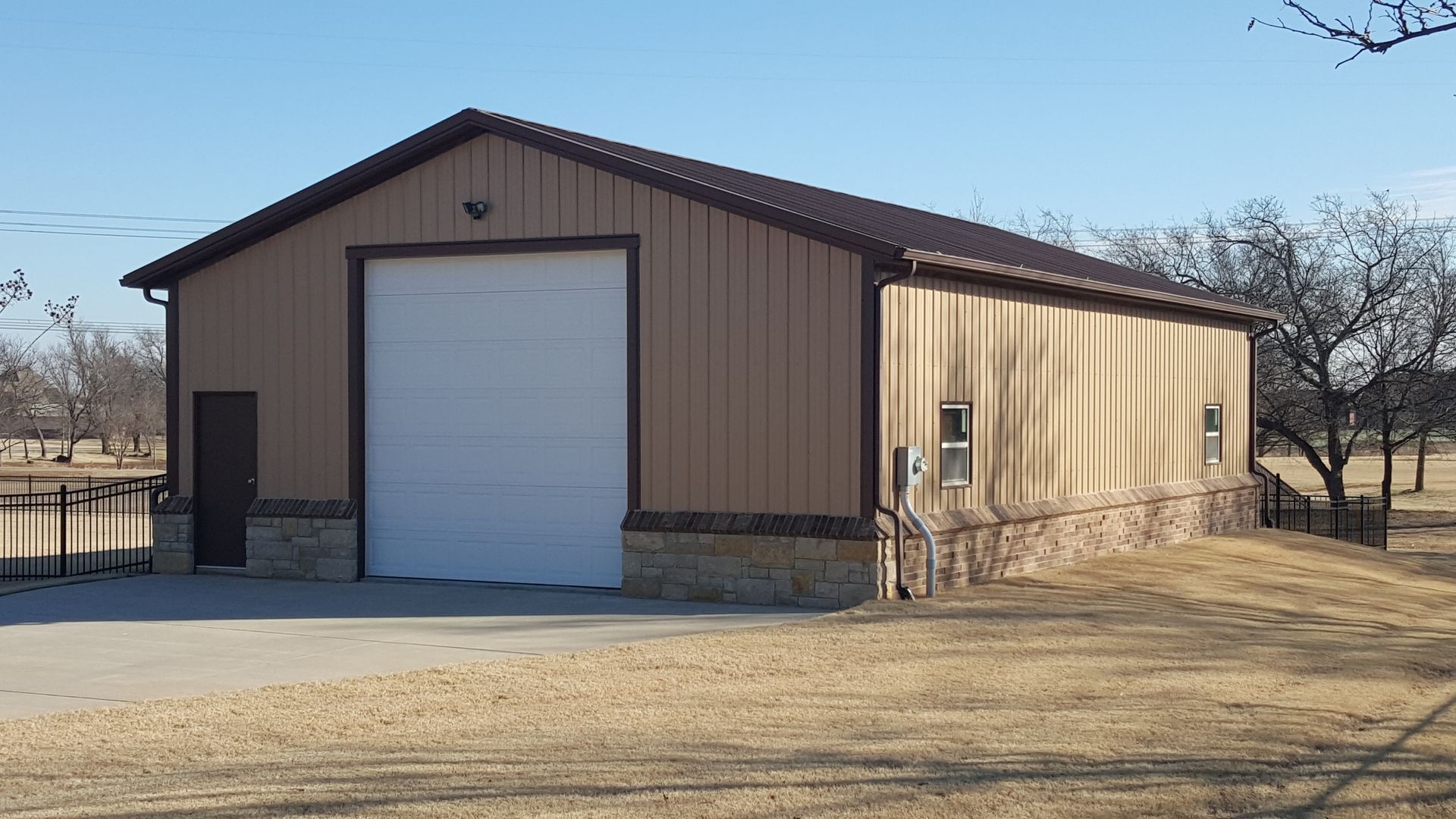 Tan metal building with a white garage door and brown roof, set in a grassy area on a sunny day.