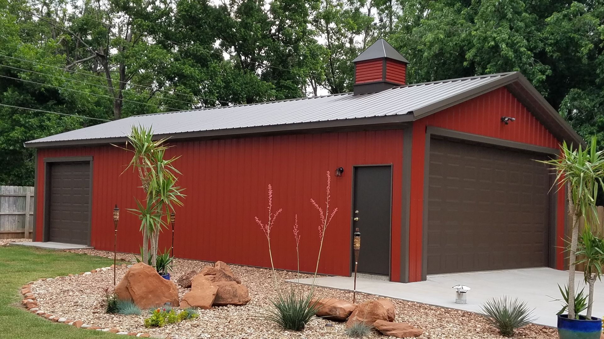 Red metal garage with brown doors and roof, in a grassy yard with a garden bed.