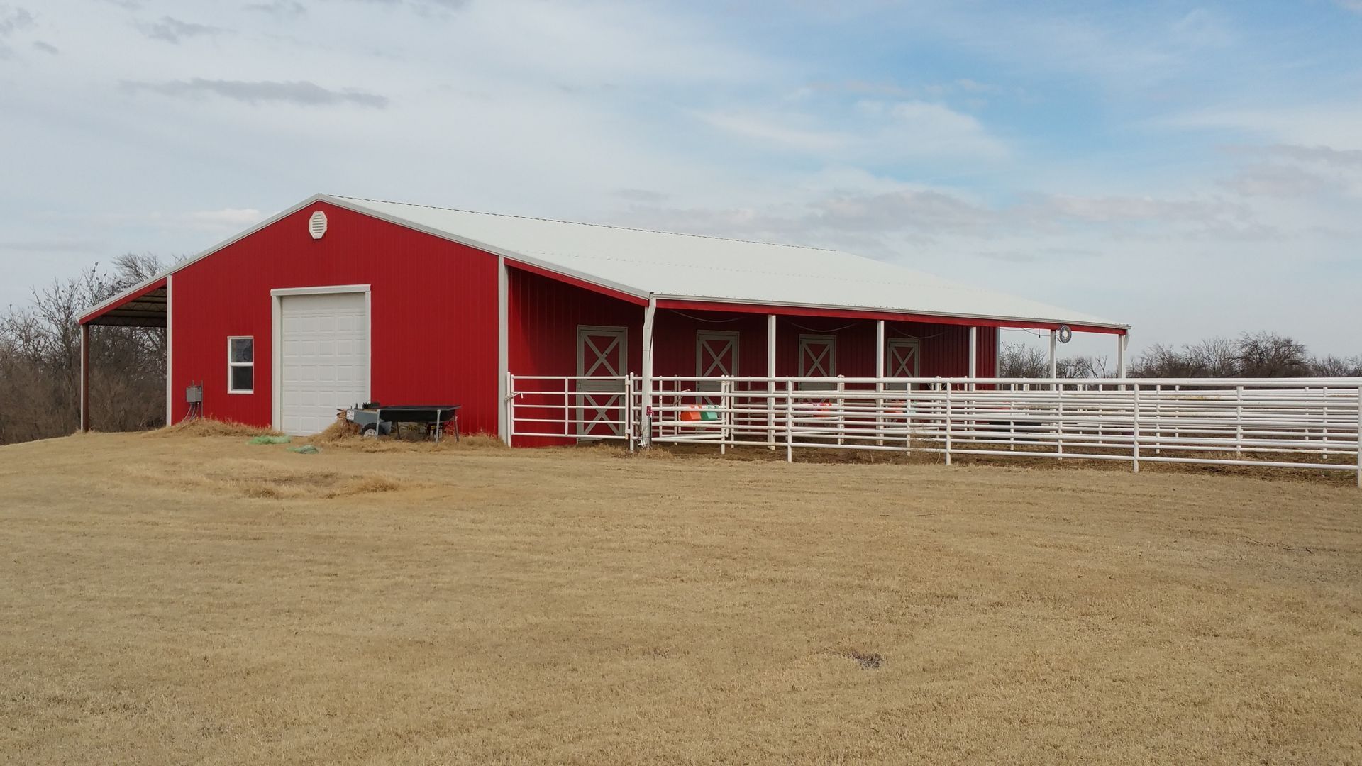 Red barn with white trim, fence, and gravel lot under a cloudy sky.