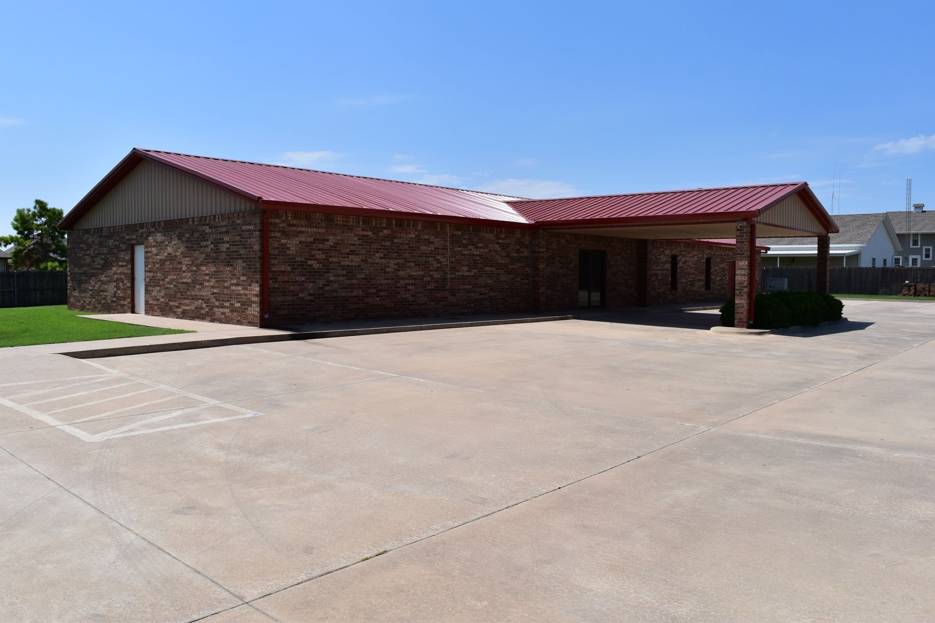 Brick building with red metal roof and a concrete parking lot on a sunny day.