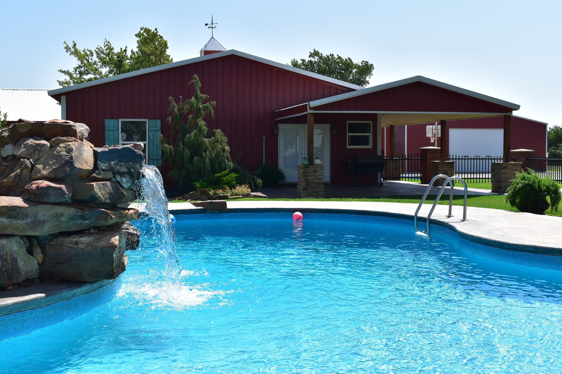 Red barn with pool, waterfall, and blue water on a sunny day.