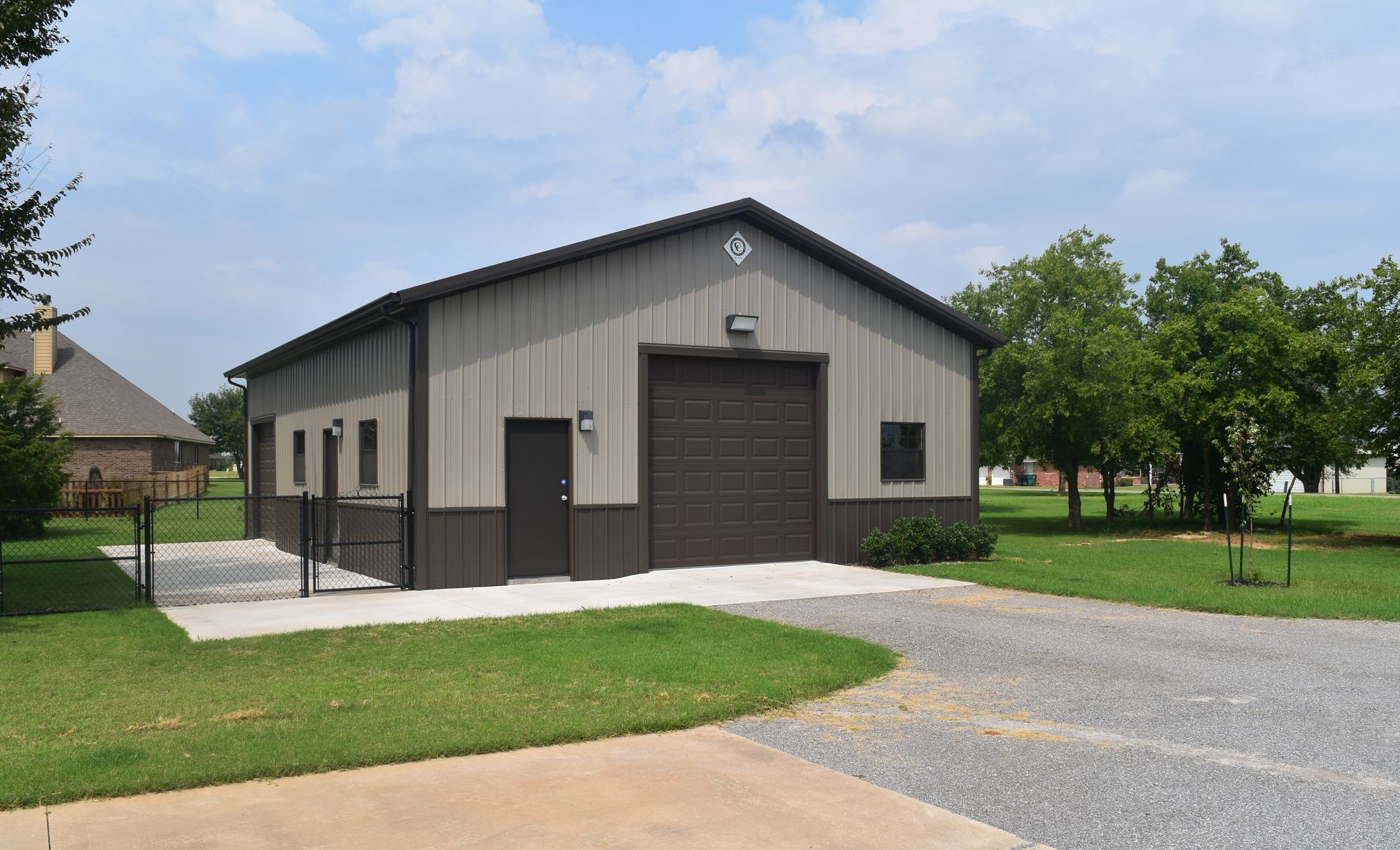 Tan and brown metal building with a driveway and green grass, under a partly cloudy sky.