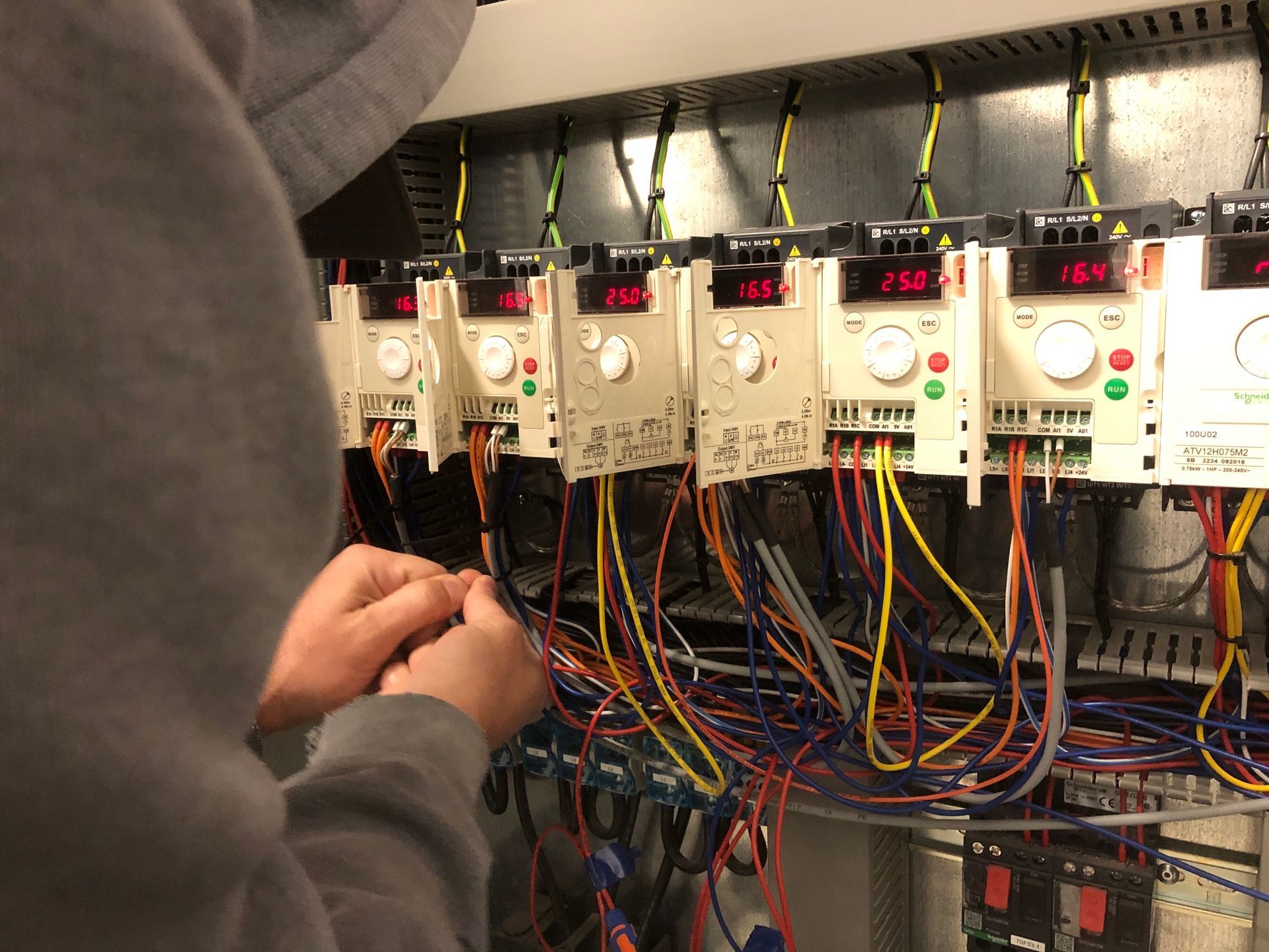 Person wiring electrical components in a control panel with multiple digital displays and colored wires.