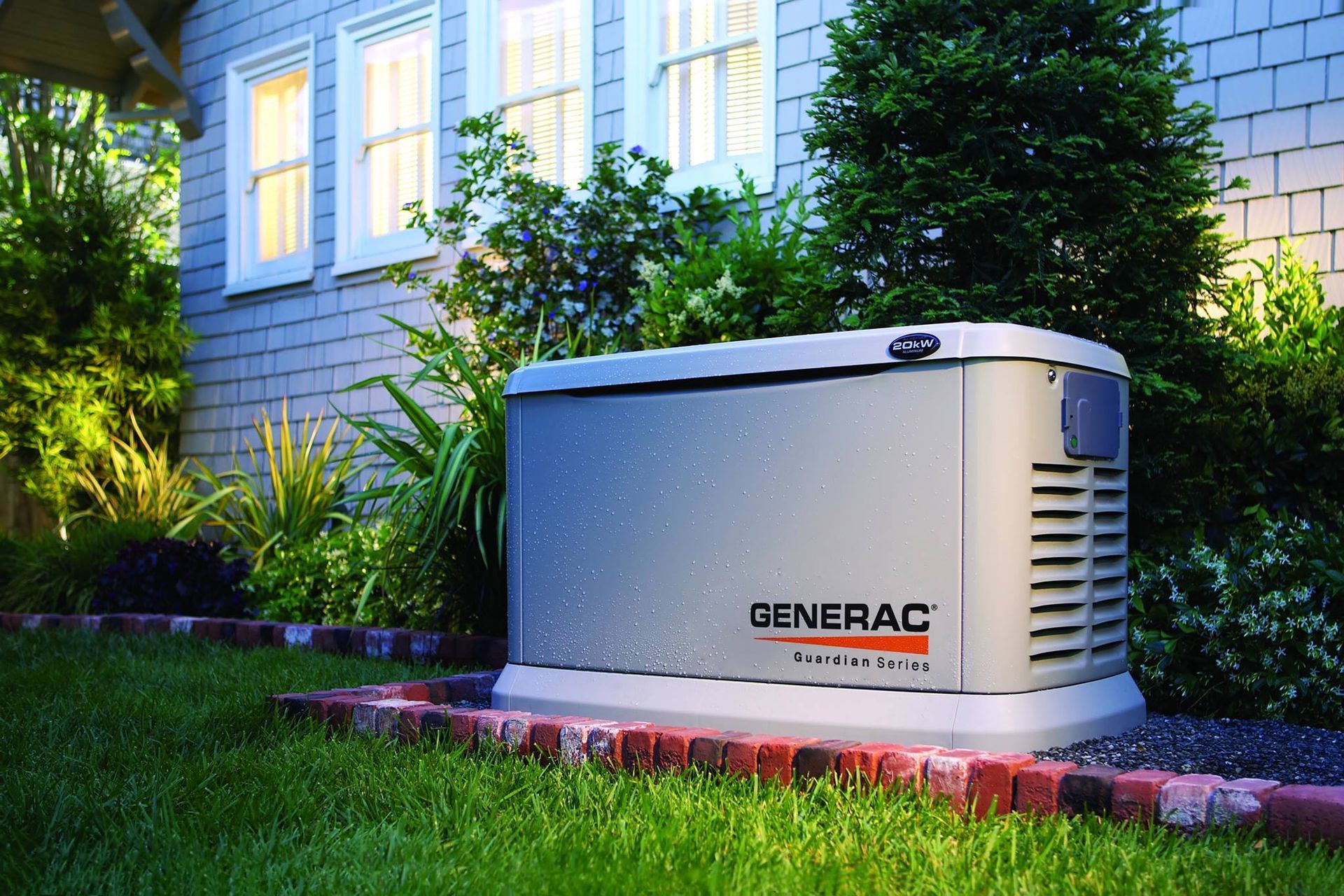 Generac home generator in a residential yard. Gray unit with brick border, next to a house with shrubs.