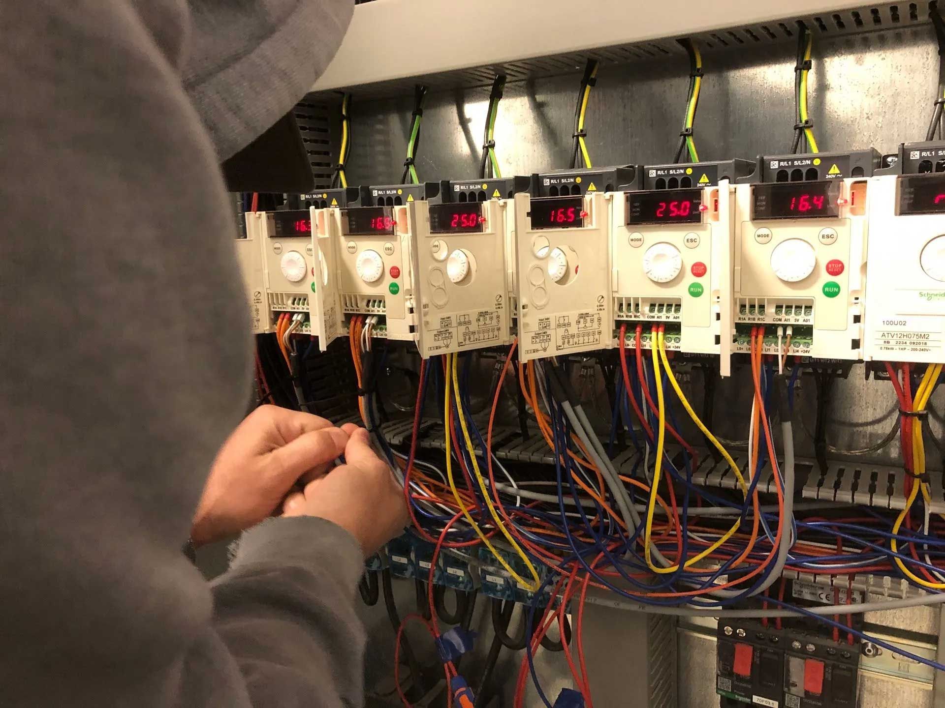 Person working with wires in an electrical panel, with multiple frequency drives displaying digital readouts.
