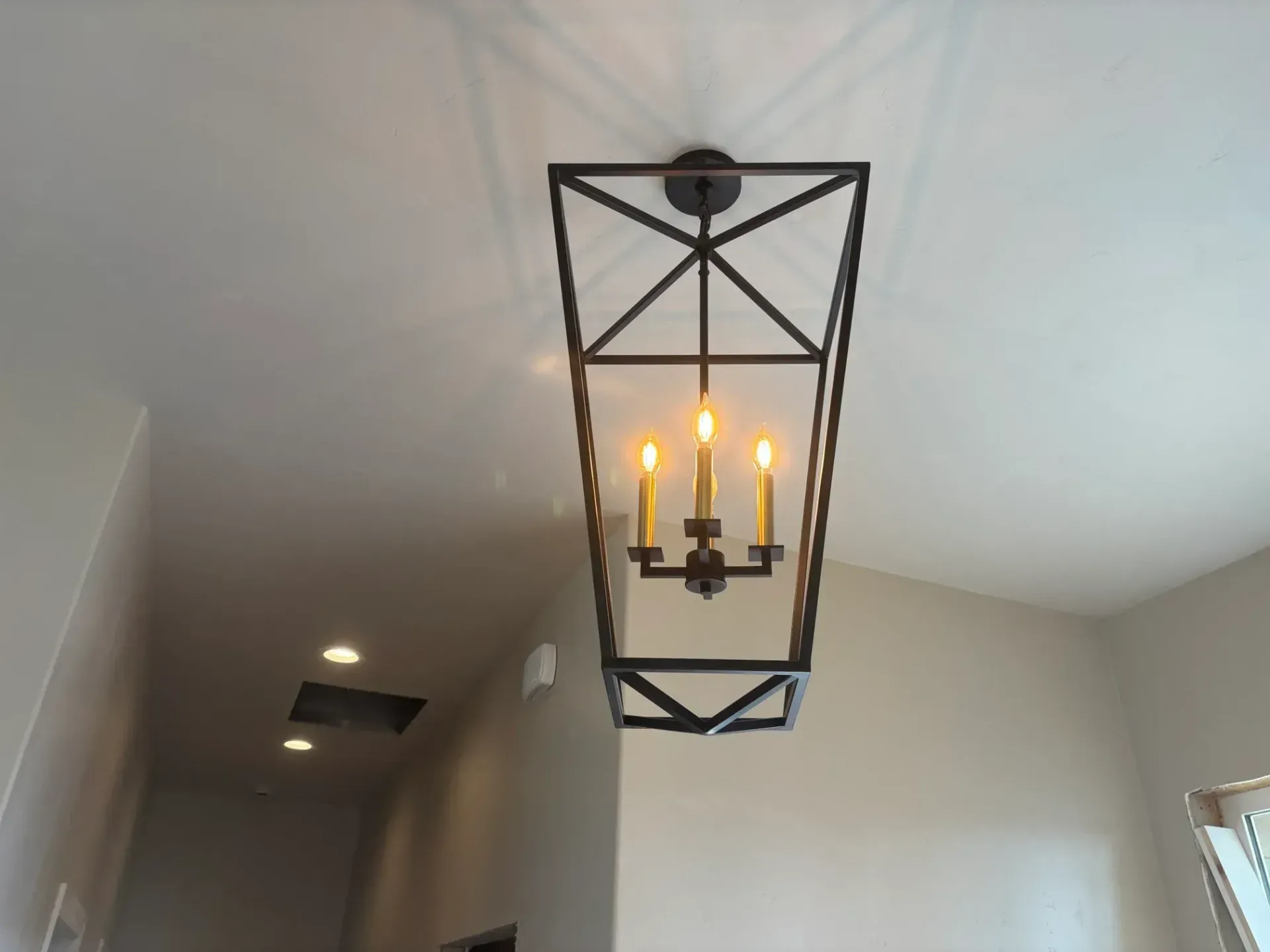Black geometric chandelier with three lit candle-shaped bulbs in a neutral-toned hallway.