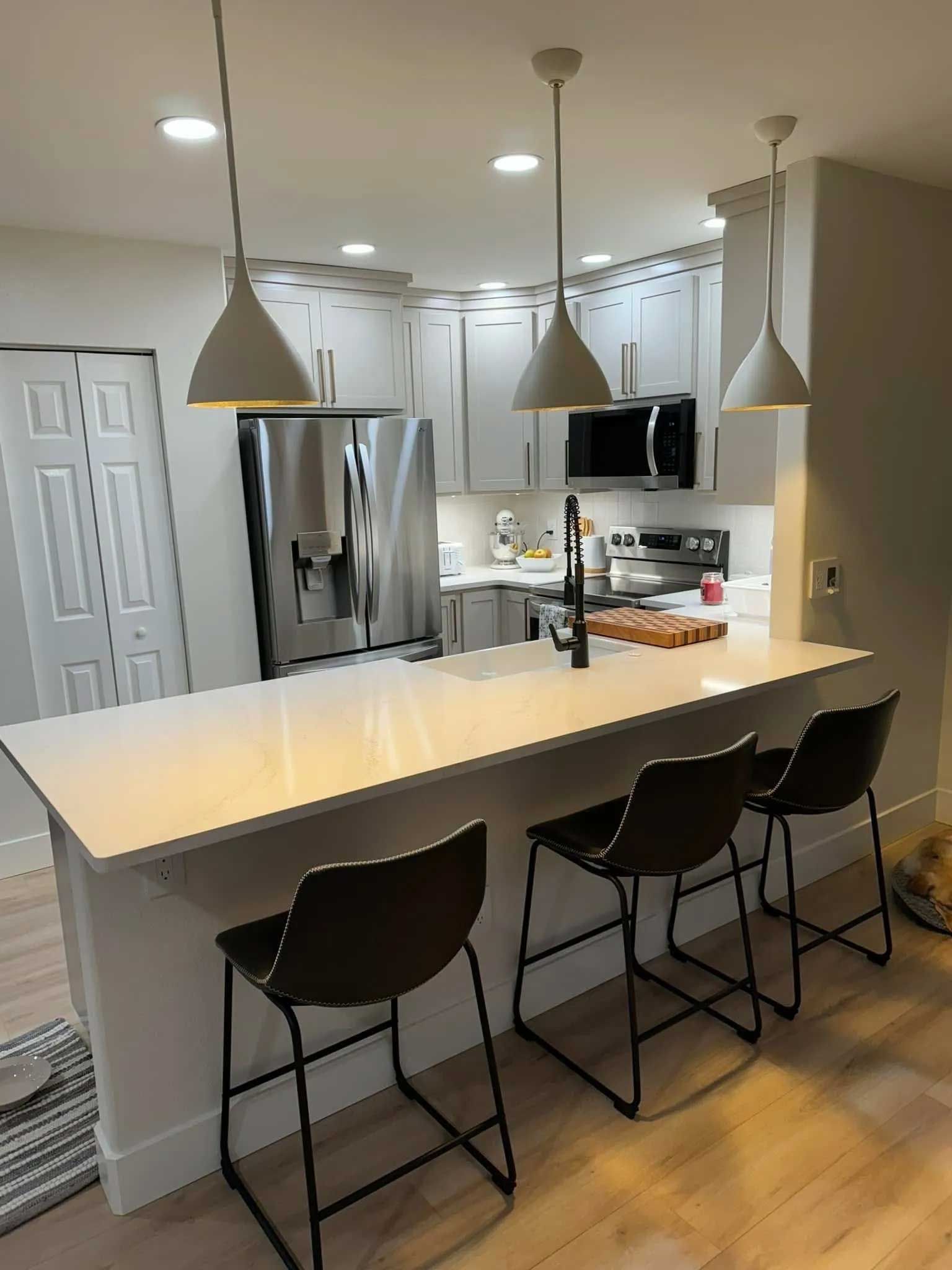 Modern kitchen with white cabinets, stainless steel appliances, breakfast bar with three black chairs, and pendant lights.