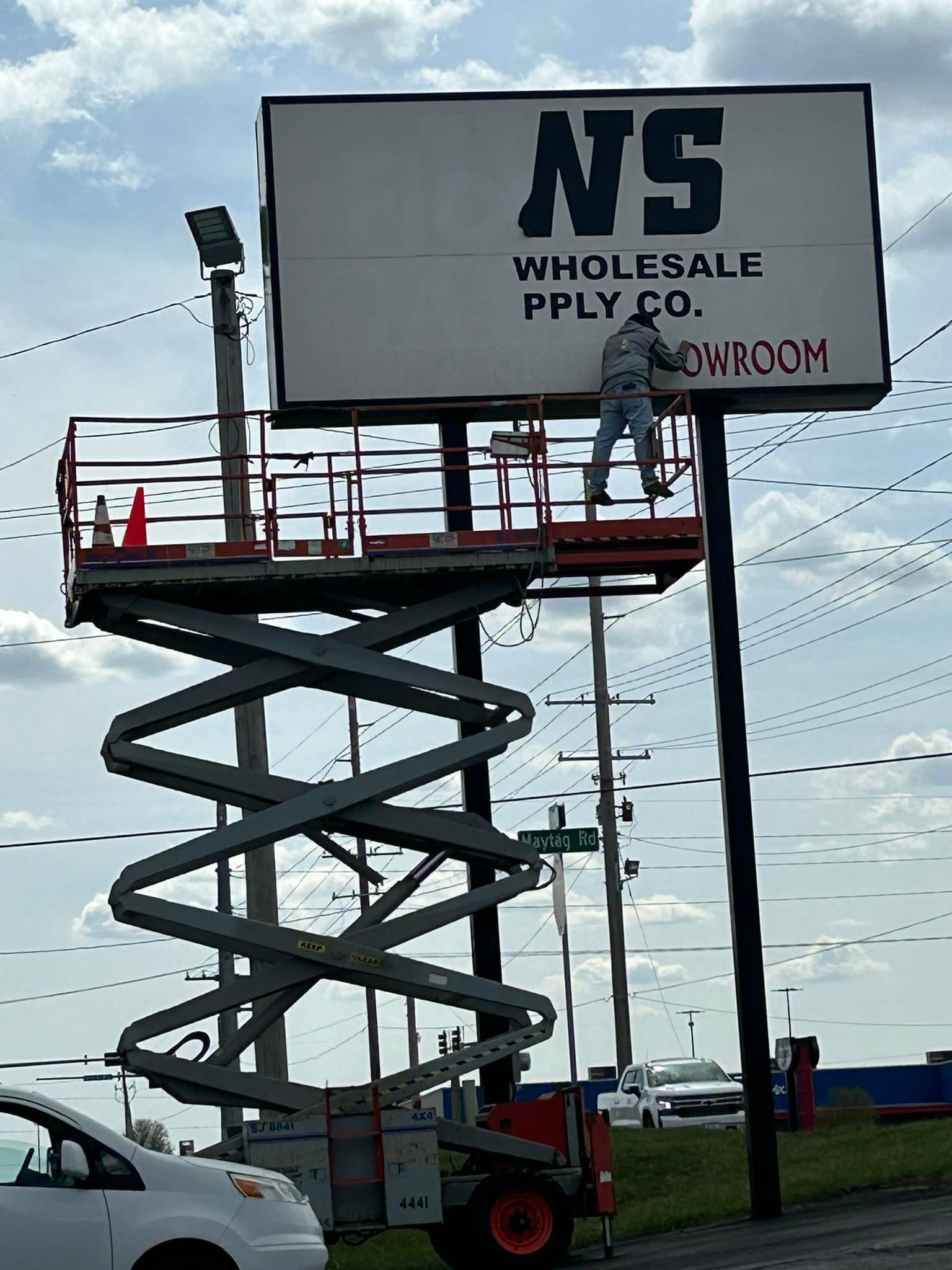 A man on a scissor lift is painting a billboard for ns wholesale pply co.