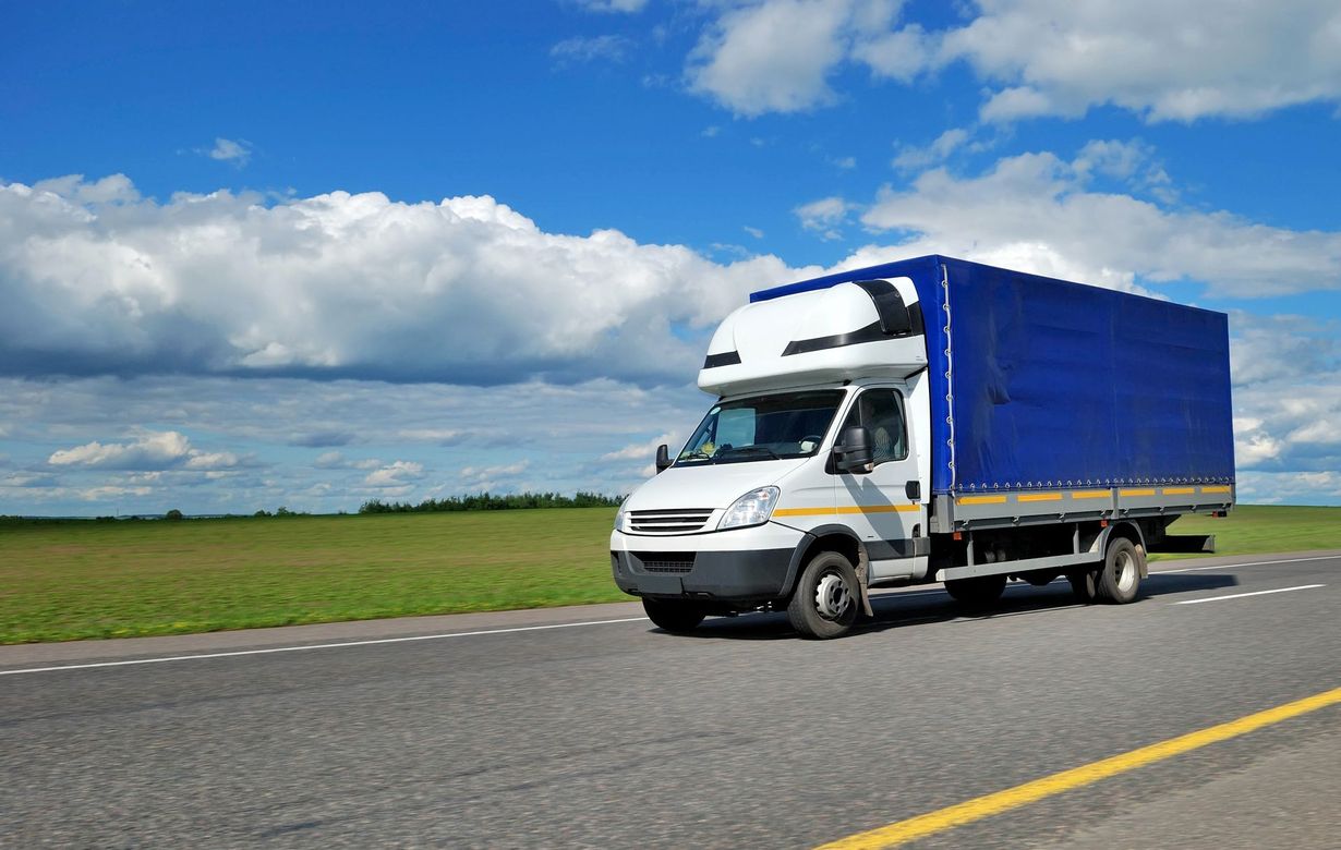White box truck with blue cargo cover driving on a rural road under a cloudy blue sky