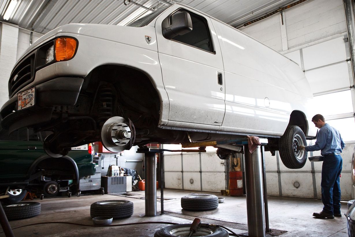 White van raised on a lift in a garage, with a mechanic standing nearby.