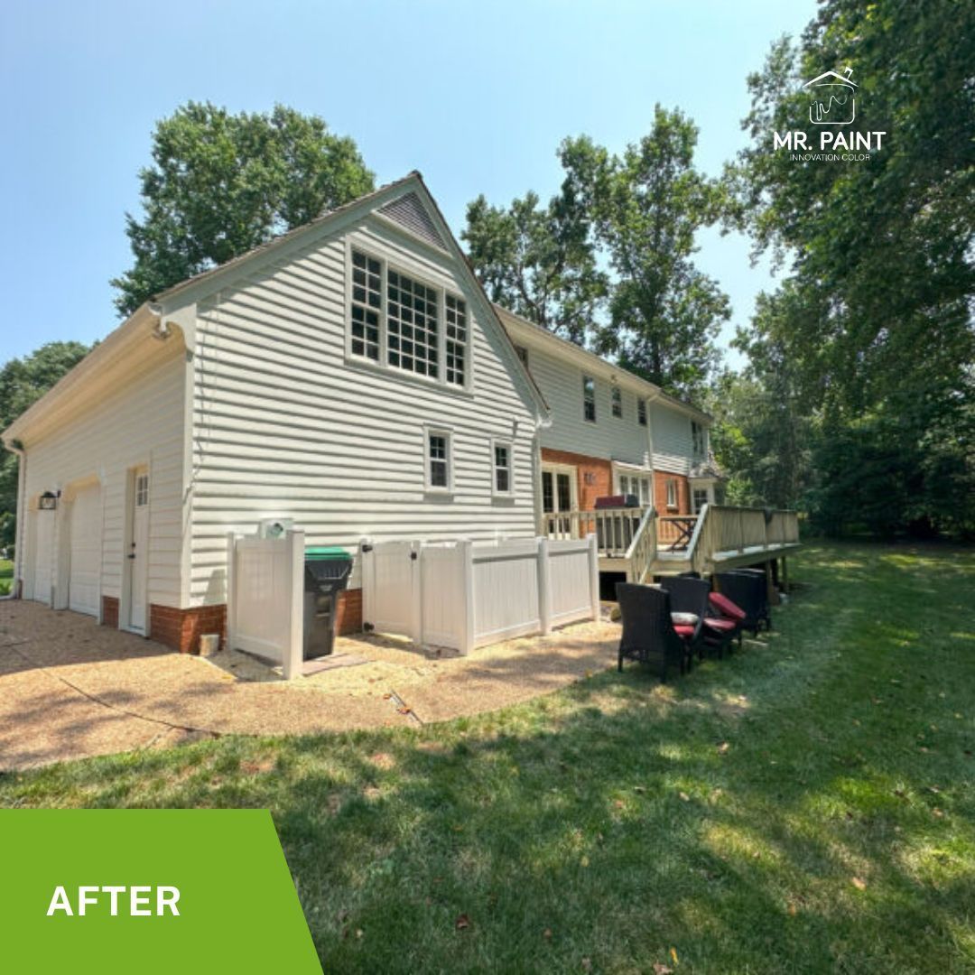 Rear view of a light-colored house with a patio and deck, surrounded by a grassy yard and trees.
