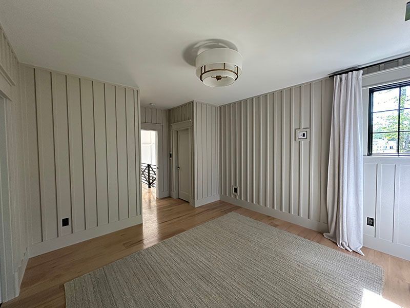 Empty room with light-colored, vertically grooved walls, light wood floor, and a rug. A white curtain hangs by a window.