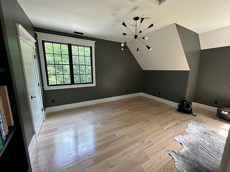 Empty room with gray walls, light wood floor, white trim, black window, and modern chandelier.