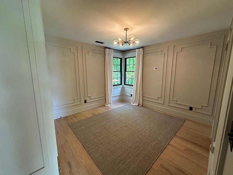 Empty room with beige trim, light wood floors, rug, and chandelier. Corner window with white curtains.