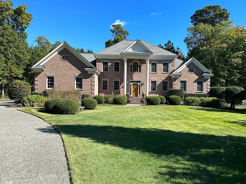 Brick two-story house with white columns, green lawn, and circular driveway. Blue sky, bushes.