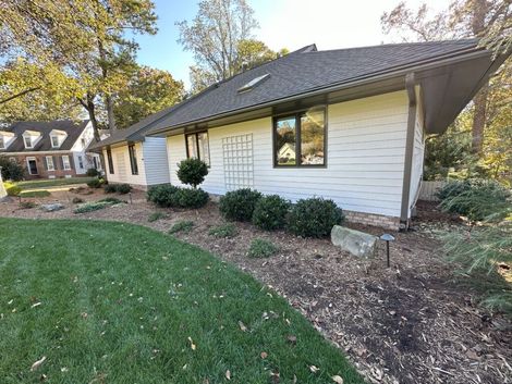 White house with angled roof, shrubs, and mulch landscaping in front. A grassy lawn and trees surround the house.