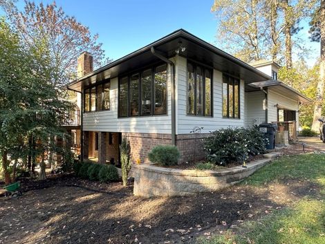 A light-colored house with dark trim and large windows sits on a slight hill, surrounded by trees and shrubs.