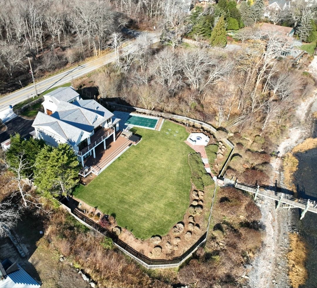 Aerial view of a large house with a pool, lawn, and a small bridge over water.