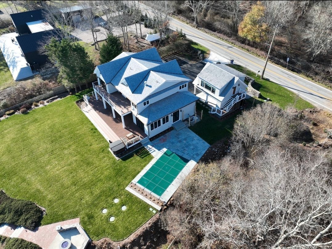 Aerial view of a light gray house with a blue pool, green lawn, and a smaller white house.