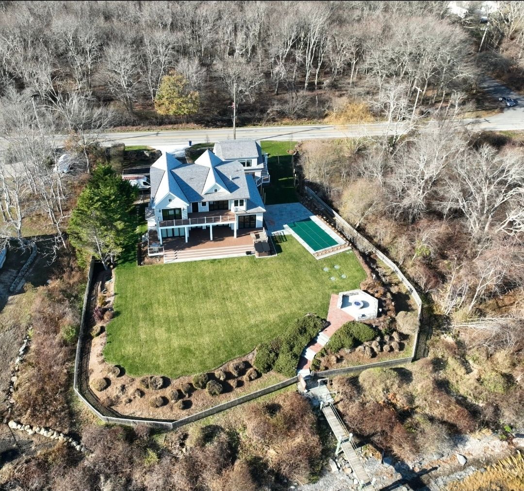 Aerial view of a large, white house with a pool and green lawn by a road and trees.
