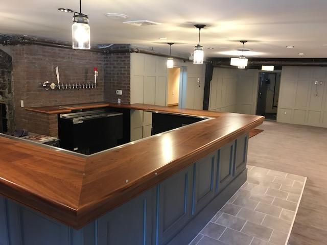A newly constructed bar with a wooden countertop, gray trim, and beer taps against a brick wall. Empty space with cabinets is in the background.