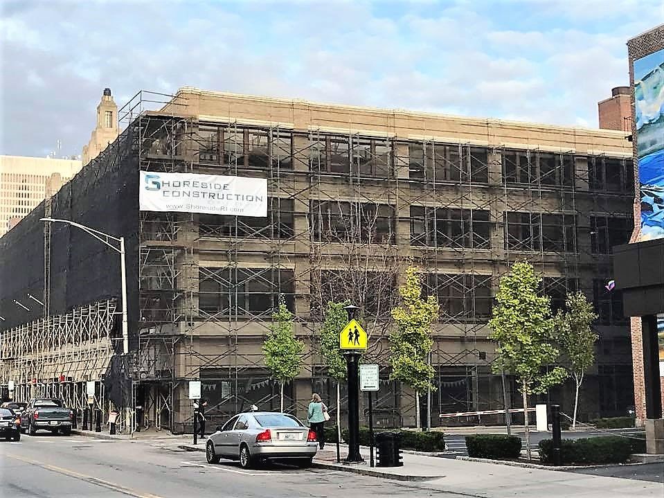 Exterior of a multi-story building under renovation with scaffolding and covered in black netting, cars and pedestrians on the street in front.