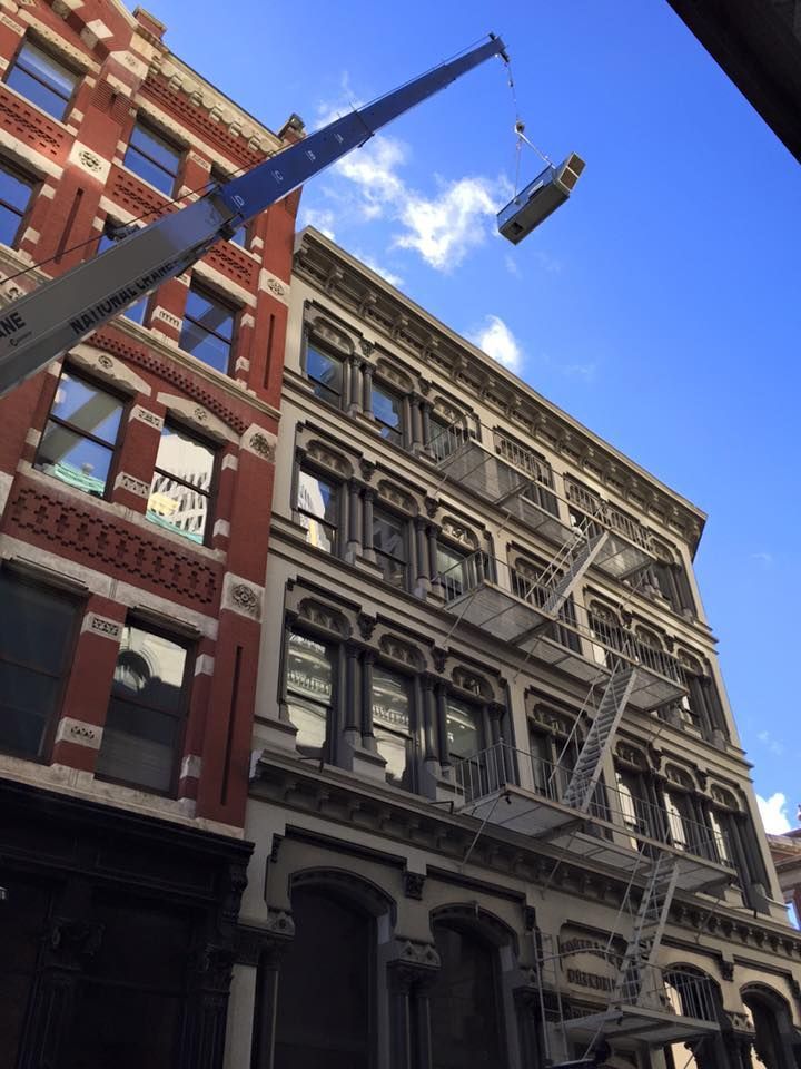A crane hoists a large object (possibly a bottle) between two brick buildings with fire escapes under a blue sky.