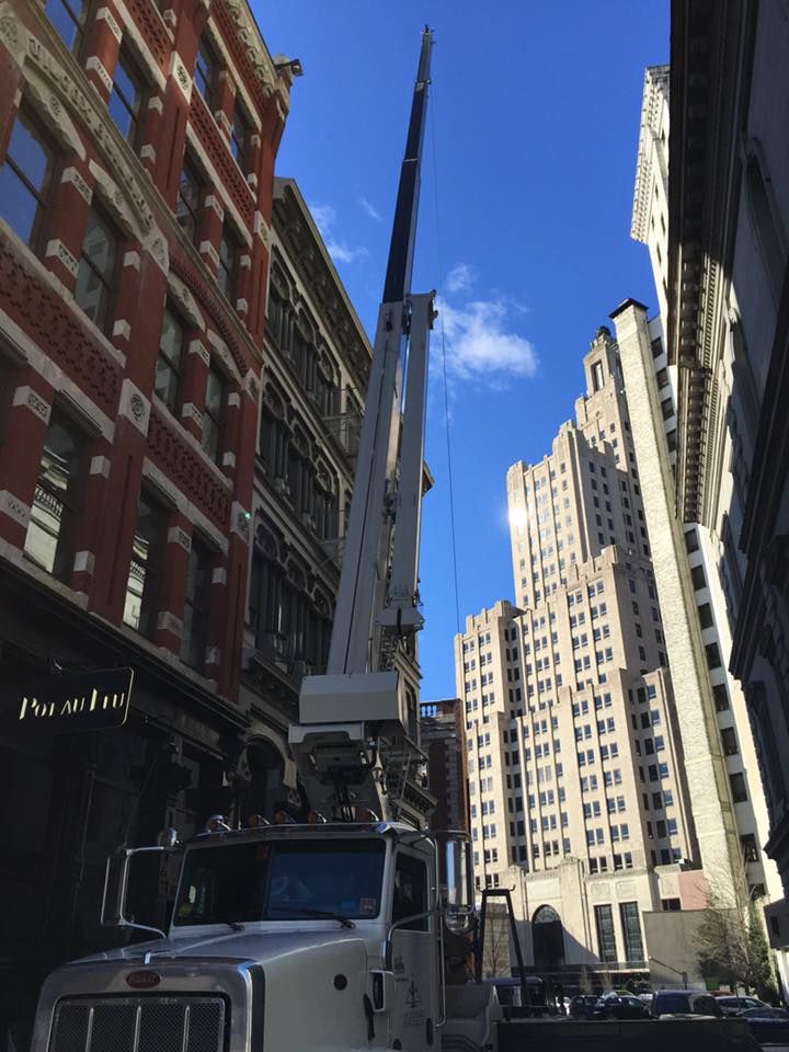 A white crane truck in a city street. Tall brick buildings flank it. Clear blue sky.