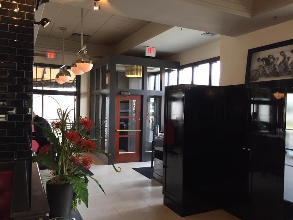 Interior of a restaurant entrance with a floral arrangement, dark cabinets, and a glass-enclosed doorway. The space is well-lit.
