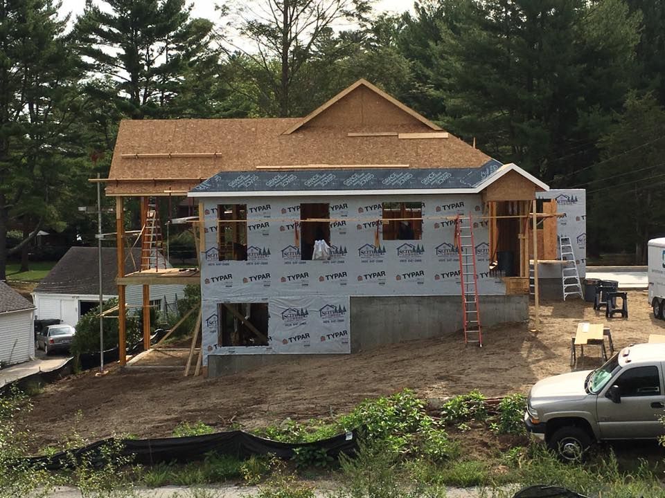 House under construction with wooden framing and sheathing. Brown roof and walls wrapped in weather-resistant material.