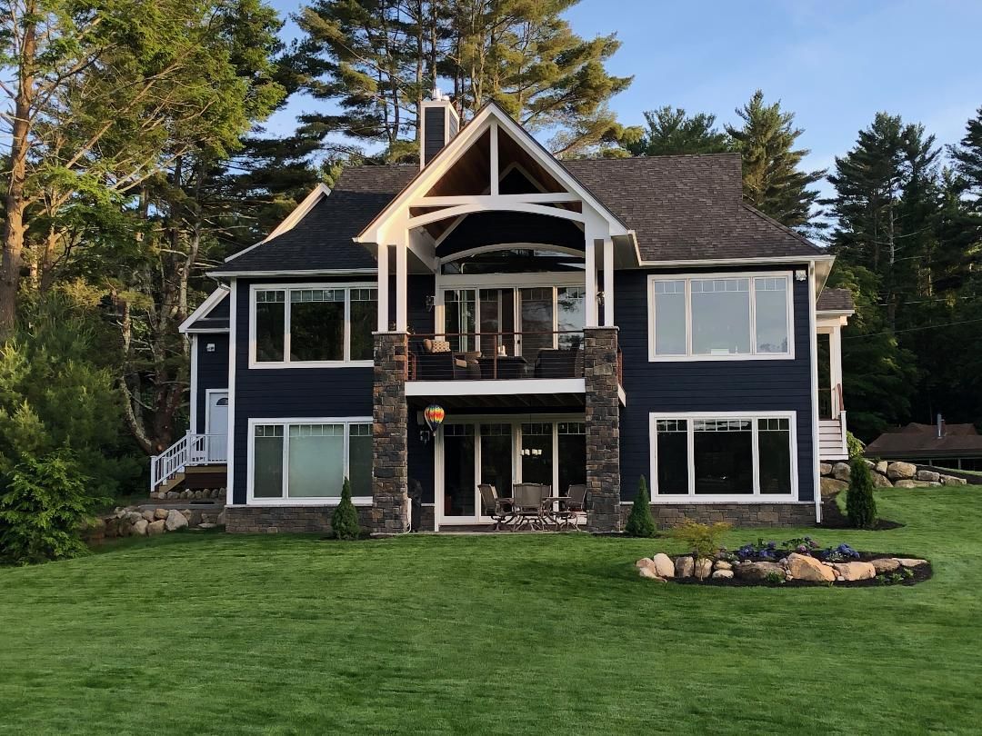 Blue house with white trim and stone accents on a green lawn by a lake with trees in the background.