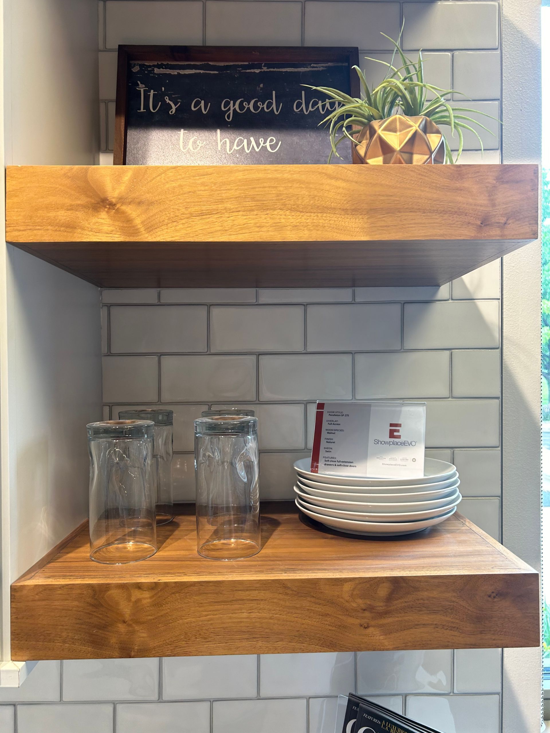 A kitchen with three wooden shelves filled with plates , glasses , and a sign.