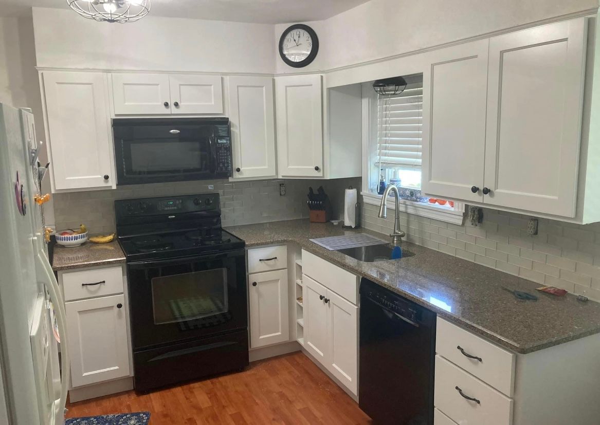 A kitchen with white cabinets , black appliances , granite counter tops and a clock on the wall.