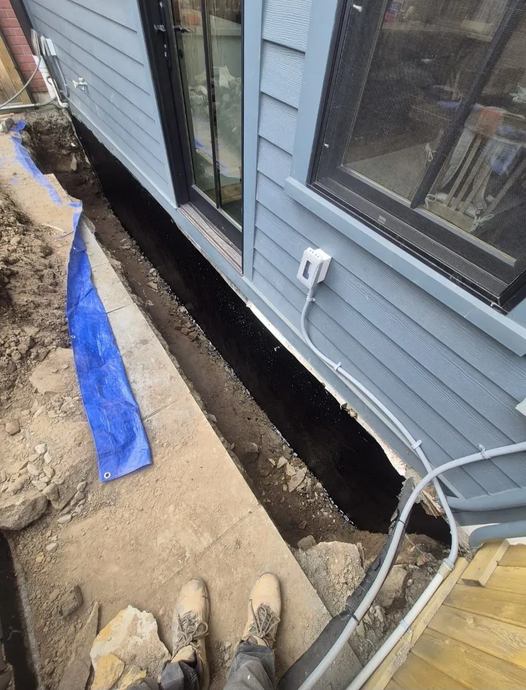 Narrow dirt trench beside blue house siding and windows, with a blue tarp and hose along the edge.