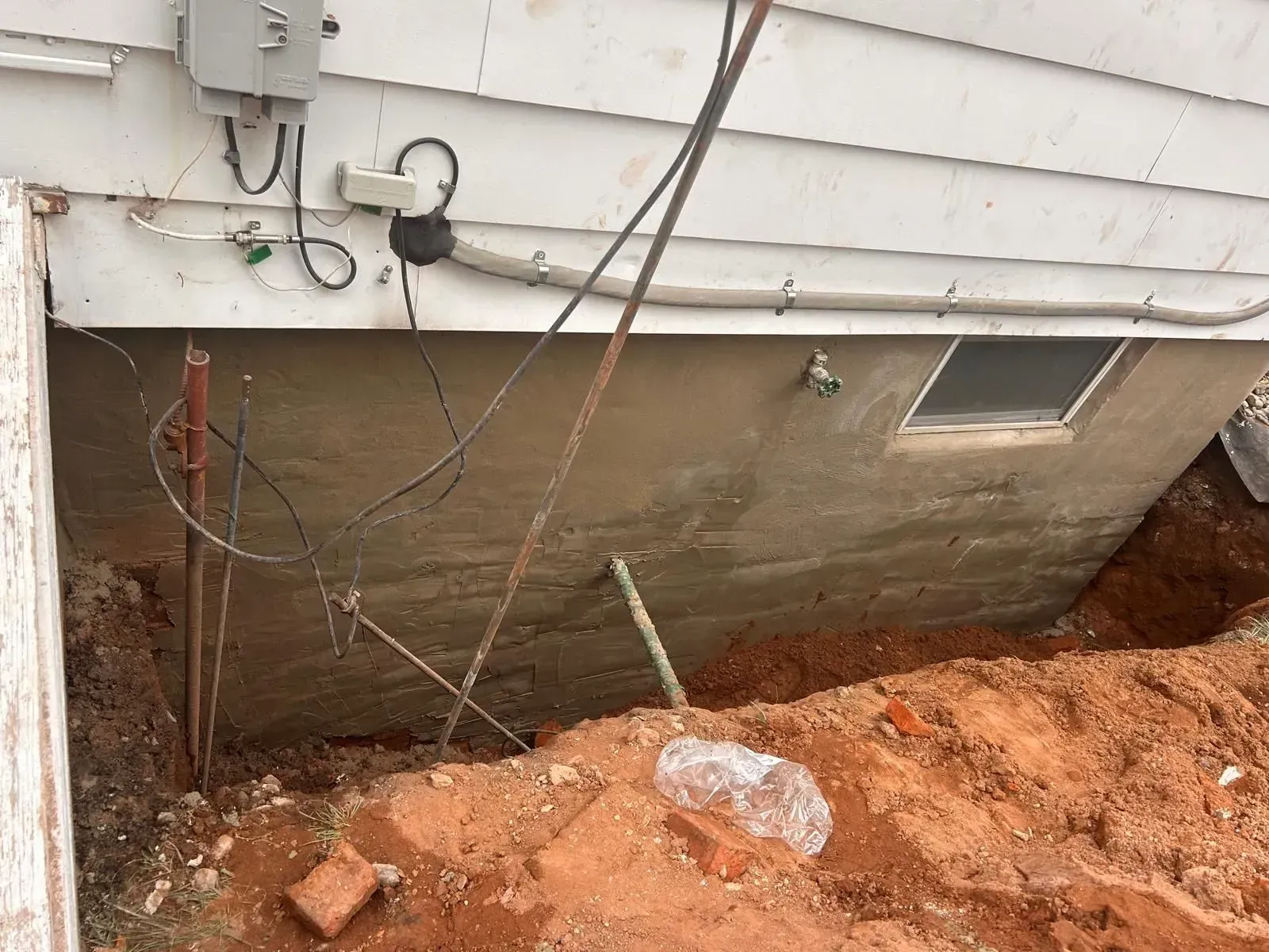 House foundation wall with exposed soil, cables, and a small basement window during excavation work
