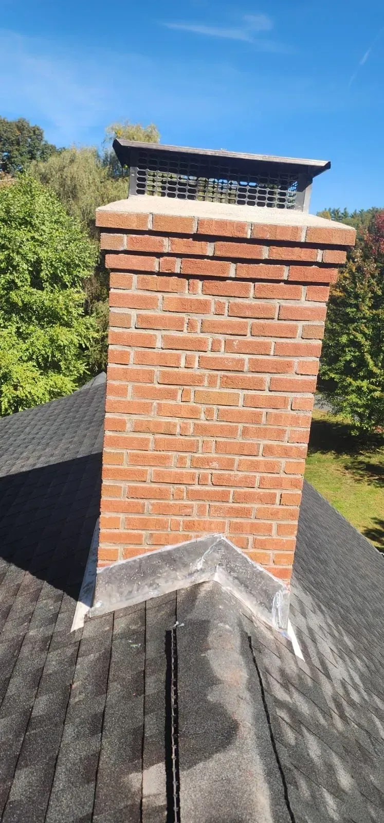 Brick chimney rising from a dark shingled roof against a blue sky and green trees