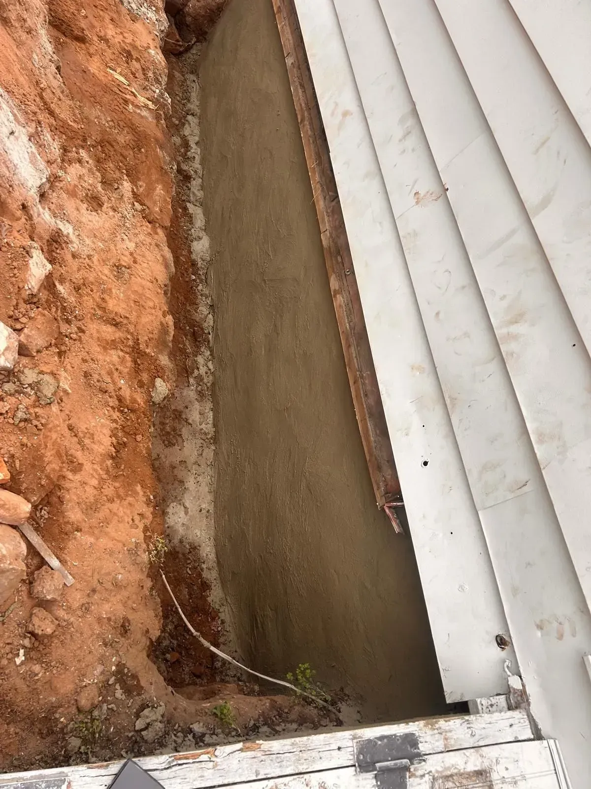 Narrow concrete drainage trench beside red dirt and white siding, with exposed wire near the bottom.