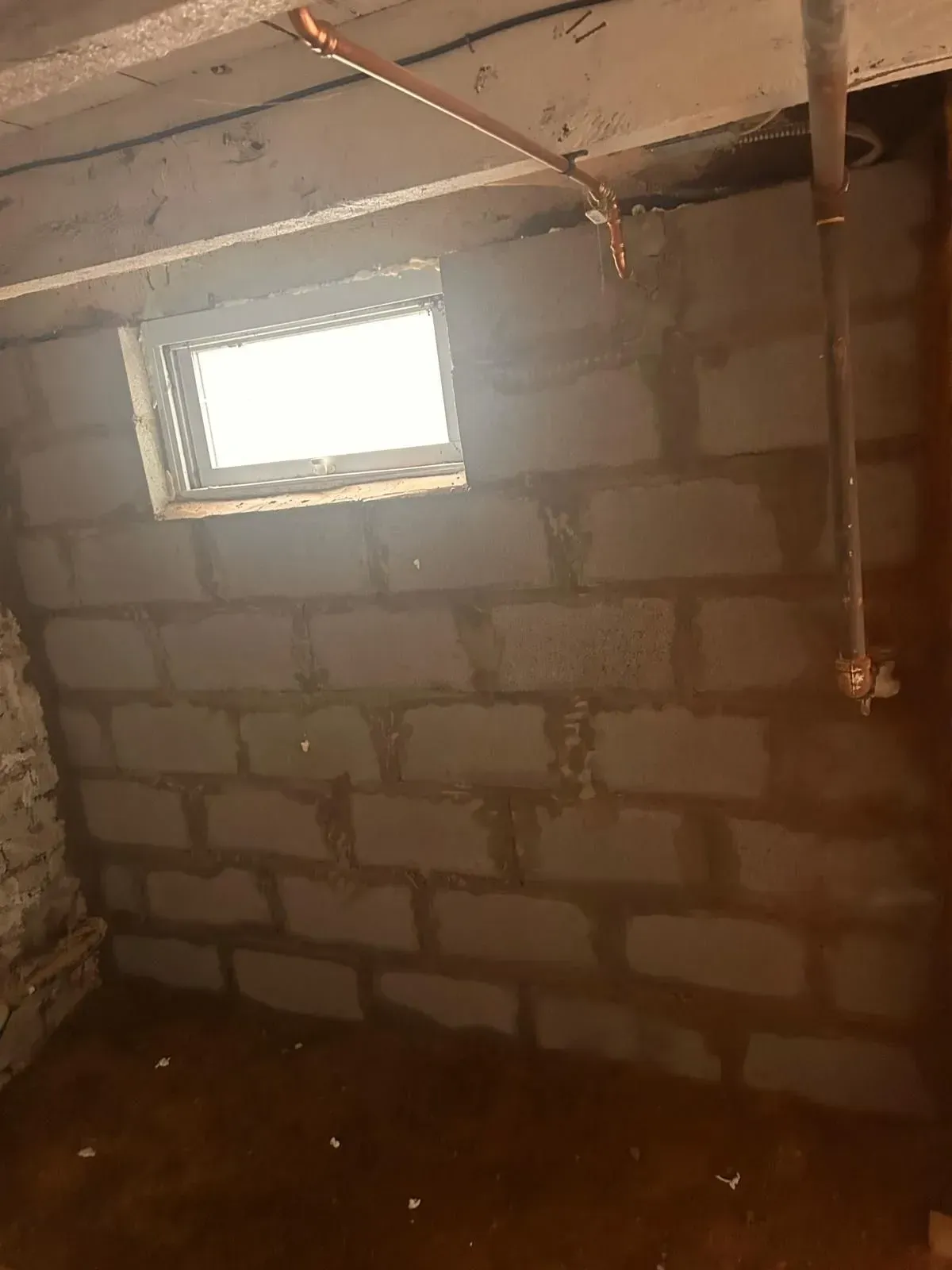 Unfinished cinder block room with a small window and exposed ceiling pipes, lit by daylight.