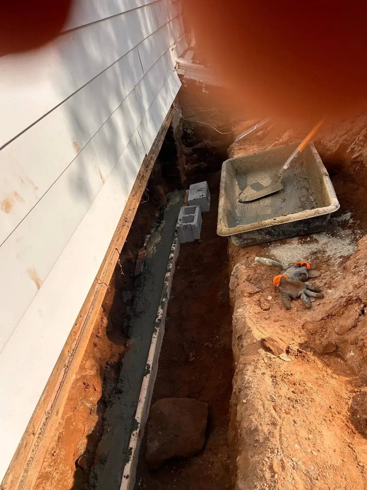 Narrow trench beside a house foundation with exposed pipes, a utility box, and dirt excavation.